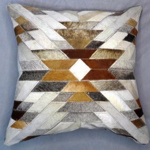 Puede incluir: Una almohada cuadrada con un patrón geométrico hecho de diferentes tonos de marrón, gris y blanco de cuero. La almohada está hecha de piel de vaca y tiene un tacto suave y lujoso.
