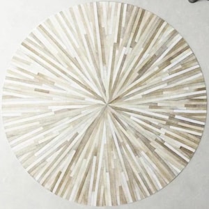 Könnte beinhalten: Ein runder, weißer und brauner Kuhfellteppich mit einem Sonnenstrahlenmuster. Der Teppich besteht aus Lederstreifen, die zusammengenäht sind, um ein einzigartiges und stilvolles Design zu schaffen.