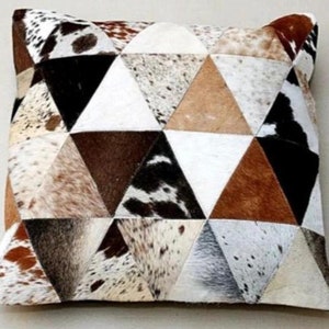 Puede incluir: Una almohada decorativa con un patrón geométrico hecho de piel de vaca. La almohada está hecha de parches triangulares de piel de vaca marrón, blanca y negra.