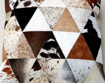May include: A decorative pillow with a geometric pattern made from cowhide. The pillow is made up of triangular patches of brown, white, and black cowhide.