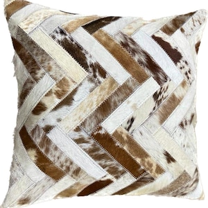 May include: A square throw pillow with a patchwork design made of brown, white, and tan cowhide. The pillow is made of various pieces of leather sewn together in a herringbone pattern.