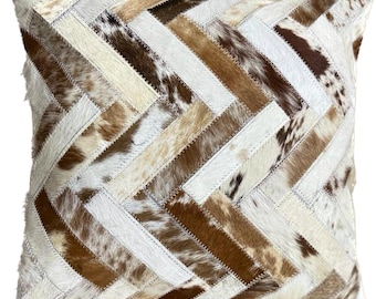 May include: A square throw pillow with a patchwork design made of brown, white, and tan cowhide. The pillow is made of various pieces of leather sewn together in a herringbone pattern.