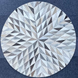 May include: A round, white and gray area rug made of leather. The rug has a geometric pattern of triangles.