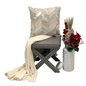 May include: A gray upholstered ottoman with a white throw blanket and a white and silver chevron patterned pillow. The ottoman has a cross-shaped base with silver studs. A white vase with red flowers sits on the floor next to the ottoman.