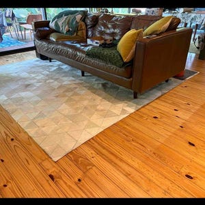 Peut inclure: Un canapé en cuir marron clair avec un tapis géométrique blanc et marron devant lui. Le canapé a plusieurs coussins, dont un coussin vert à motif floral, un coussin jaune et un coussin marron.