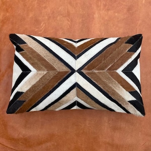 May include: A rectangular pillow with a geometric pattern made of brown, black, and white cowhide. The pillow is on a brown leather surface.