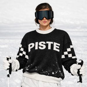 Könnte beinhalten: Eine Person trägt einen schwarzen Pullover mit dem Wort "PISTE" in Weiß. Der Pullover hat weiß-schwarze Karomuster auf den Ärmeln. Die Person trägt eine Skibrille und hält Skistöcke.