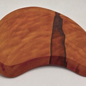 May include: A handcrafted wooden massage tool with a smooth, curved design. The wood has a warm, reddish-brown hue with a dark, vertical grain pattern. The tool is designed to fit comfortably in the hand for massage.