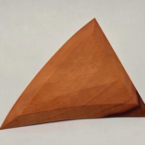 May include: A triangular, wooden object with a warm, reddish-brown hue. The geometric shape has a flat base and a curved top, with visible wood grain. The object appears to be hand-carved and has a smooth finish.