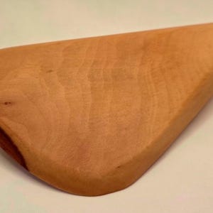 May include: A wooden cutting board with a unique, asymmetrical shape. The board is made of light brown wood with a darker, reddish-brown accent on one corner. The wood grain is visible, adding to its natural aesthetic.