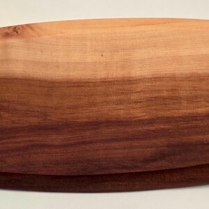 May include: Oval-shaped wooden serving board with a natural wood grain finish. The board features a two-tone design, with a lighter brown top and a darker brown base. The edges are rounded, and the board appears to be made of a solid piece of wood.