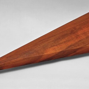 May include: A handcrafted, triangular wooden object with a sharp point. The wood has a rich, reddish-brown color and visible grain patterns. The object appears to be a decorative piece or a functional item, possibly a serving utensil or a decorative element.