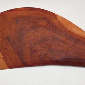 May include: A handcrafted wooden massage tool with a teardrop shape. The tool is made of two-toned wood, with a lighter, tan section and a darker, reddish-brown section. The wood grain is visible.