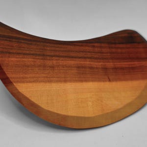 May include: A wooden, teardrop-shaped cutting board with a natural wood grain pattern. The board features a gradient of colors, transitioning from light tan to dark brown. The curved design adds a unique aesthetic to this kitchen item.