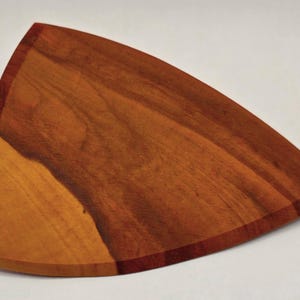 May include: A wooden serving board with a unique, curved triangular shape. The board is made of a warm-toned wood, with a rich, reddish-brown border. The grain of the wood is visible, adding to its natural aesthetic.