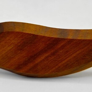 May include: A handcrafted wooden bowl with a flowing, organic shape. The bowl is made of a rich, dark brown wood with visible grain patterns. The design is reminiscent of a leaf or a flame, with a smooth, polished finish.