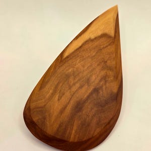 May include: A teardrop-shaped wooden object with a smooth, polished surface. The wood grain is visible, showcasing a blend of light and dark brown tones. The object has a rounded base and a pointed tip.