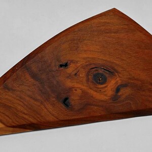 May include: A handcrafted, triangular wooden cutting board with a rich, dark brown color and natural wood grain patterns. The board features a unique knot design and a beveled edge, showcasing the natural beauty of the wood.