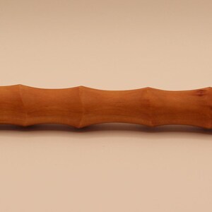 May include: A wooden massage roller, crafted from a warm, medium brown wood. The roller features a series of rounded ridges along its length, and the ends are slightly wider. The design gives it a bamboo-like appearance. The wood grain is visible.