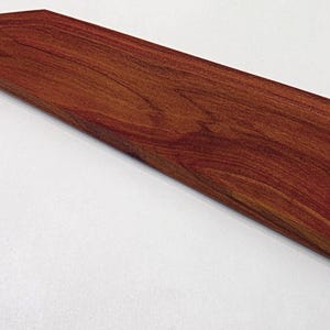 Pode incluir: Um objeto de madeira longo e angulado, com uma rica cor castanho-avermelhada e grão de madeira visível. A peça tem uma forma geométrica, com um topo plano e lados angulados, e parece ser um item decorativo ou funcional, possivelmente uma tábua de cortar ou para servir.