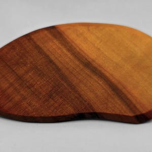 May include: A wooden, organic-shaped cutting board with a rich, warm color gradient. The board transitions from a deep, reddish-brown to a lighter, golden hue, showcasing the natural wood grain. The board is approximately 10 inches long.