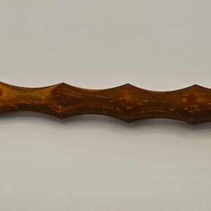 May include: A dark brown wooden massage stick. The stick has a series of curved ridges along its length, designed for muscle relief. The wood grain is visible. The massage stick is approximately 25 cm long.