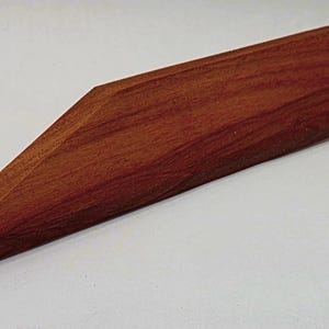 Puede incluir: Un tope de puerta de madera hecho a mano con un rico acabado marrón rojizo. El tope de puerta tiene una parte superior rectangular que se estrecha hasta un punto afilado, creando una forma de cuña. La veta de la madera es visible.