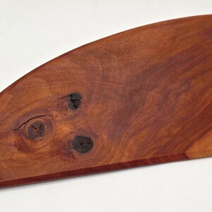 Puede incluir: Tabla de cortar de madera hecha a mano con una forma curva única. La tabla está hecha de madera de color marrón rojizo, con vetas y nudos naturales. La tabla mide aproximadamente 30 cm de largo y 20 cm de ancho.