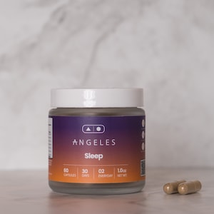 Può includere: Un barattolo di vetro con un coperchio bianco che contiene capsule marroni. Il barattolo è etichettato "ANGELES Sleep" e presenta un design sfumato in viola, arancione e giallo. L'etichetta indica anche "60 capsule, 30 giorni, 02 ogni giorno, 1,0 oz peso netto."