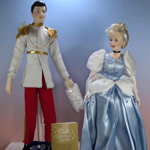 May include: Two porcelain dolls representing a prince and Cinderella. The prince wears a grey jacket with gold epaulets and red trousers. Cinderella wears a light blue satin gown with white puff sleeves. Includes a glass slipper, a booklet, and a card.