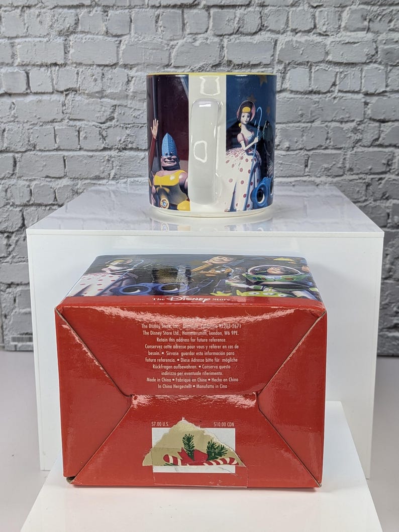 May include: A ceramic mug featuring Toy Story characters including Bo Peep and a blue-helmeted figure, paired with a red cardboard box. The box base displays The Disney Store text, manufacturing details, and price labels of $7.00 US and $10.00 CDN.