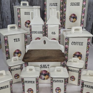 May include: A collection of white ceramic kitchen canisters and bottles featuring black text labels and floral borders. The set includes containers for flour, sugar, rice, tea, coffee, salt, pepper, nutmeg, allspice, cloves, ginger, and cinnamon.