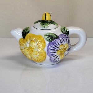 May include: A small, white ceramic teapot with a lid and handle. The teapot is decorated with yellow and purple flower designs and green leaves. The lid has a yellow accent. A charming addition to any tea set.