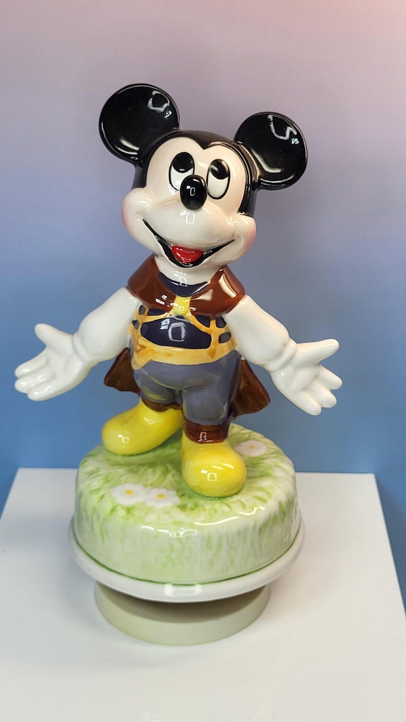 May include: A ceramic Mickey Mouse figurine with outstretched arms. Mickey is wearing a brown vest, blue pants, and yellow shoes. The figurine is on a green base with white flowers.