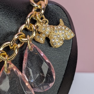 May include: A gold-toned chain bracelet with a charm featuring a rhinestone-studded dog and two pink teardrop-shaped pendants. The bracelet is displayed on a black surface against a pink background.