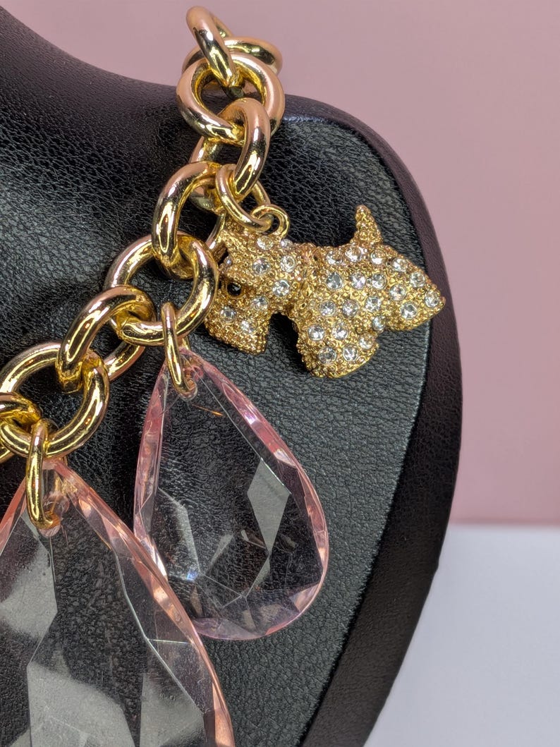 May include: A gold-toned chain bracelet with a charm featuring a rhinestone-studded dog and two pink teardrop-shaped pendants. The bracelet is displayed on a black surface against a pink background.