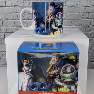 May include: A ceramic mug and its matching blue cardboard box featuring Toy Story 2 characters including Woody, Buzz Lightyear, Bo Peep, Rex, and Slinky Dog. The box displays the text Toy Story 2, One Mug, and The Disney Store logo.