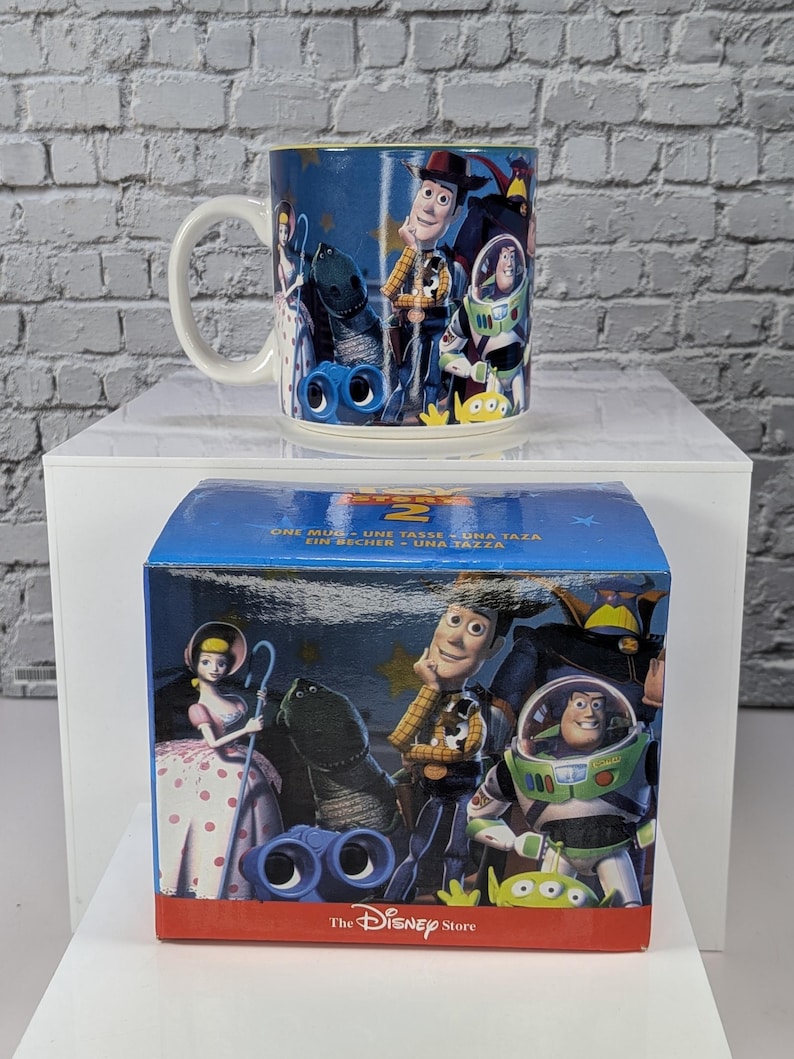 May include: A ceramic mug and its matching blue cardboard box featuring Toy Story 2 characters including Woody, Buzz Lightyear, Bo Peep, Rex, and Slinky Dog. The box displays the text Toy Story 2, One Mug, and The Disney Store logo.