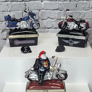 May include: Three decorative Harley-Davidson motorcycle-themed trinket boxes. One box features a Santa Claus figurine on a motorcycle. The other two boxes have motorcycle toppers in blue and burgundy. Each box has a black base with silver accents.