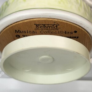 May include: A close-up of a Schmid Musical Collectibles figurine base. The base is made of wood and has the brand name printed on it. The figurine has a white and light green base. The bottom of the figurine is white.
