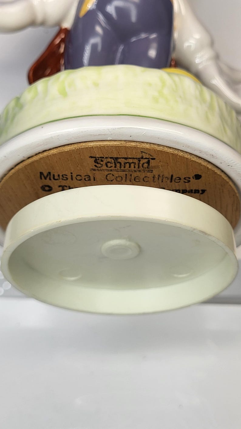 May include: A close-up of a Schmid Musical Collectibles figurine base. The base is made of wood and has the brand name printed on it. The figurine has a white and light green base. The bottom of the figurine is white.