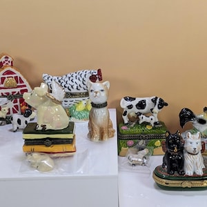 May include: A collection of ten small, hand-painted porcelain trinket boxes featuring various animal and farm-themed designs, including a red barn, a pig on books, a cat, a cow, a rooster, a duck, and several small dog figurines.