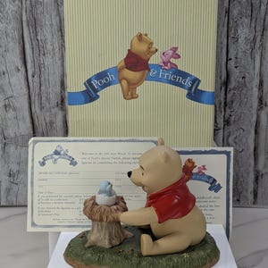 May include: A Winnie the Pooh figurine, featuring Pooh Bear in a red shirt, reaching towards a small blue bird in a nest. The base is green with a tree stump. The box behind it says "Pooh & Friends".