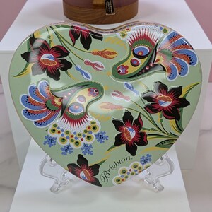 May include: Heart-shaped decorative tray with a floral design in shades of green, red, blue, and black. The tray has the word "Brighton" printed on it. The tray is displayed on a clear stand.
