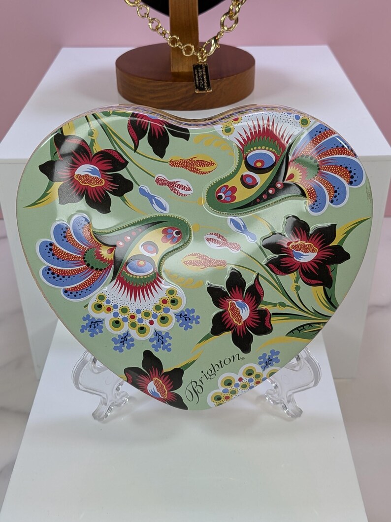 May include: Heart-shaped decorative tray with a floral design in shades of green, red, blue, and black. The tray has the word "Brighton" printed on it. The tray is displayed on a clear stand.