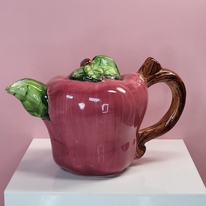 May include: A whimsical, ceramic teapot shaped like a red apple, with a brown handle and a green leaf on the lid and spout. The teapot is displayed on a white surface against a pink background.