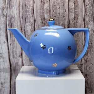 May include: A light blue ceramic teapot with a gold-colored handle and accents. The teapot features a spherical body, a spout, and a lid with a gold knob. Gold star-shaped designs are scattered across the surface. The teapot is displayed on a white surface.