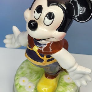 May include: A ceramic Mickey Mouse figurine with a black head and ears, white gloves, and yellow shoes. Mickey is wearing a brown vest and blue shorts, standing on a green base with white flowers.