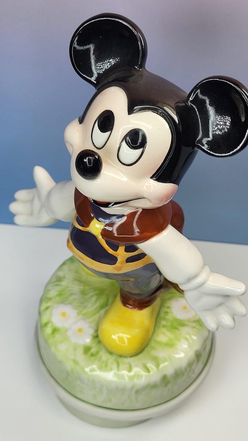 May include: A ceramic Mickey Mouse figurine with a black head and ears, white gloves, and yellow shoes. Mickey is wearing a brown vest and blue shorts, standing on a green base with white flowers.