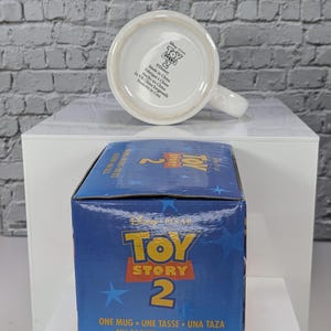 May include: A white ceramic mug with a handle, positioned behind its blue cardboard packaging box. The mug base features the Toy Story 2 logo and manufacturing text. The box displays the Toy Story 2 logo, blue stars, and the text One Mug.